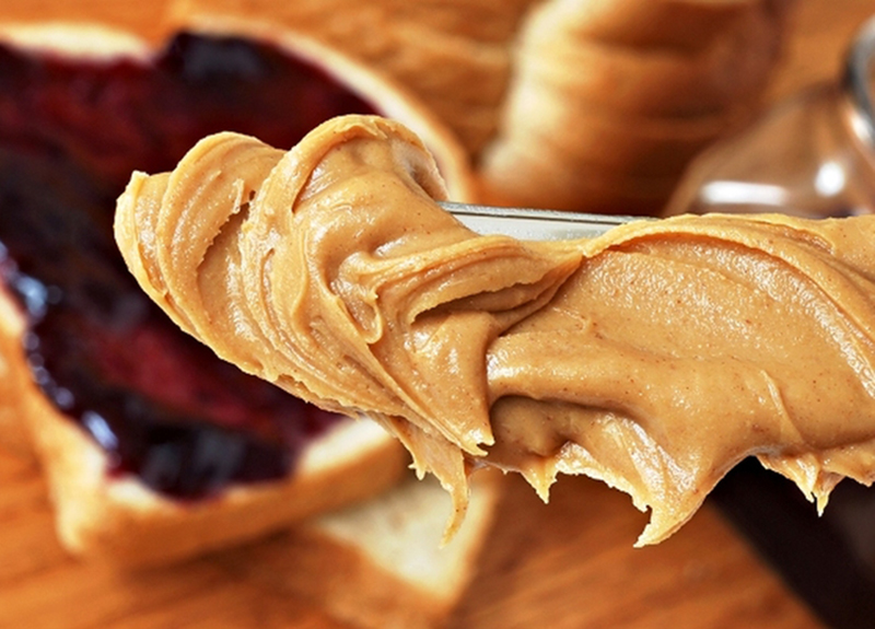 Peanut butter is spread on a knife above a PB&J sandwich.